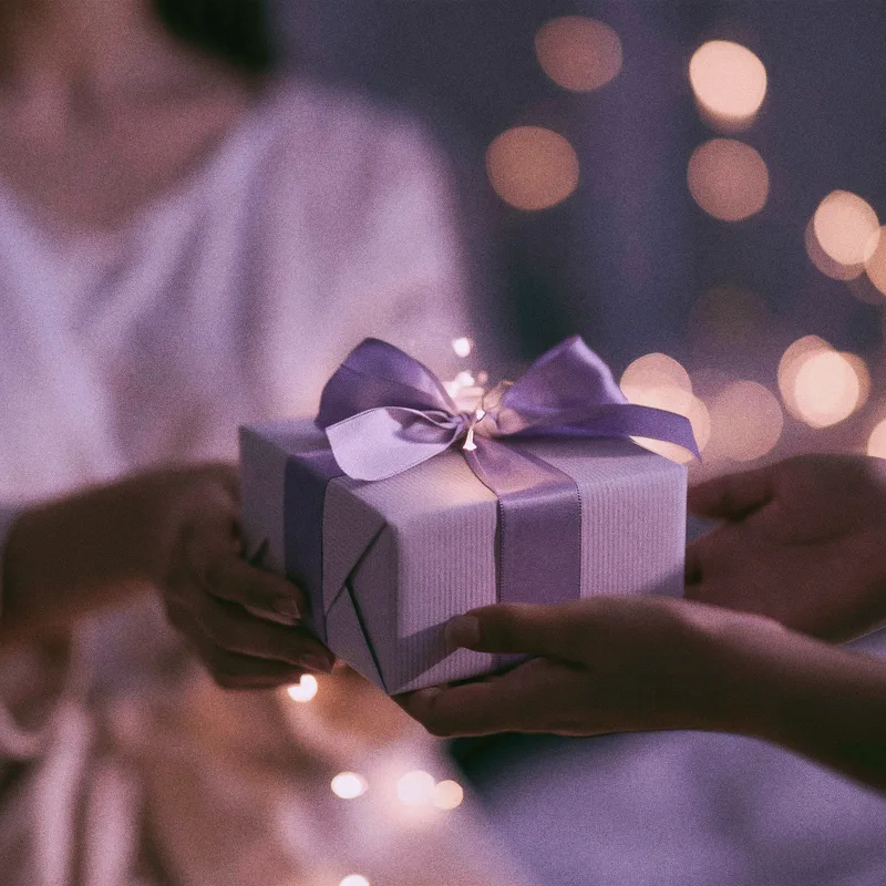 Two people passing a lilac gift box tied with a violet ribbon