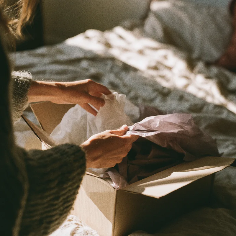 Hands folding back lilac tissue paper in an opened parcel on a bed in morning light